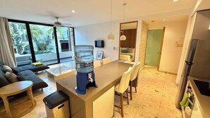 Un salon moderne comprenant un canapé, une table à manger, un distributeur d’eau et un coin cuisine avec un réfrigérateur.