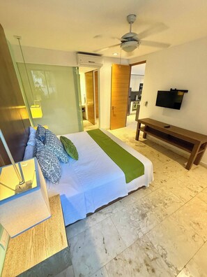 A hotel room with a bed, a TV, a ceiling fan, and a view of the kitchen.