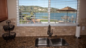 A kitchen with a double sink, a window with blinds, and a view of a lake and outdoor seating area. - Direct Lake Front Paradise Cove #1 In Reviews - Beach - Launch - Traeger Smoker (Clearlake)