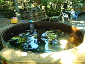 Un bain à remous en bois, agrémenté d’un banc et de chaises disposés autour, installé dans un jardin avec des arbres et une terrasse en bois.