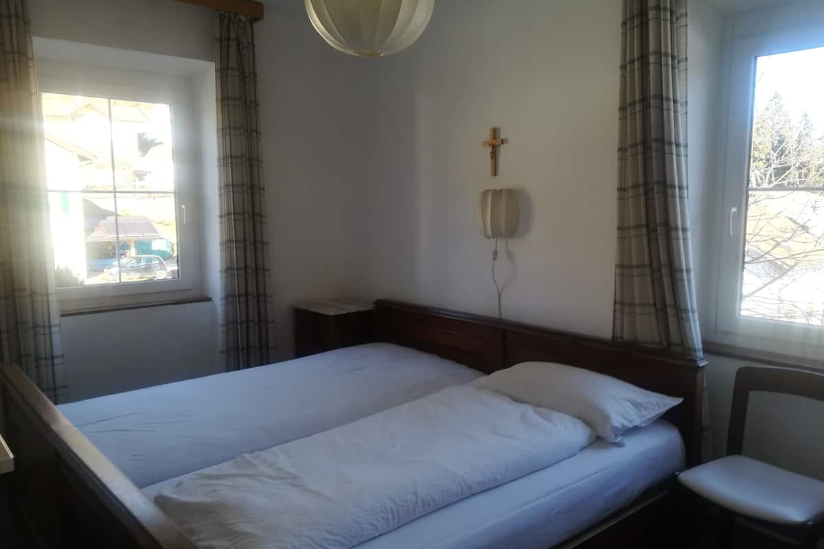 A single-bed room with a wooden headboard, a bedside table, a chair, and a window with curtains.