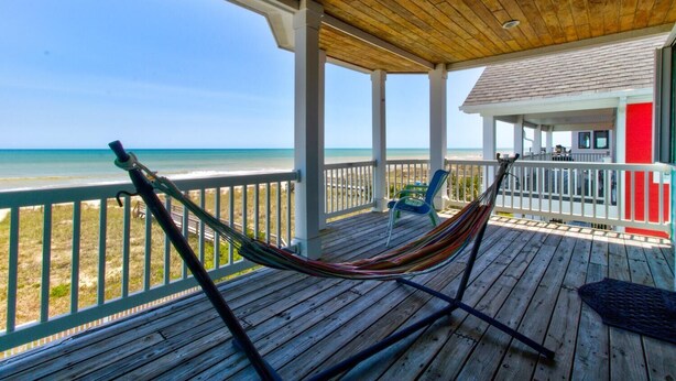 A wooden deck with a hammock, overlooking a beach and a house with a red accent.