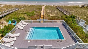 A pool area with a view of the beach, deck chairs, and a wooden deck.