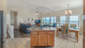 A spacious kitchen with a central island, a dining area with wooden chairs, a television, and a view of the ocean through large windows. - Callalily | Gorgeous Ocean Views in Topsail Beach! (Topsail Beach)