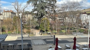 Un área de café al aire libre con mesas y sombrillas rojas, un paso de peatones y un árbol grande al fondo.