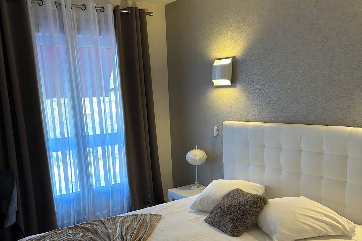 Un lit bien fait, avec une grande couverture à motifs, une tête de lit blanche et une lampe de chevet.