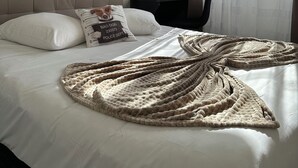 Una cama bien hecha con un edredón grande y texturizado, un cabecero, un cojín con diseño de perro, una lámpara de noche y vistas al exterior a través de cortinas traslúcidas.