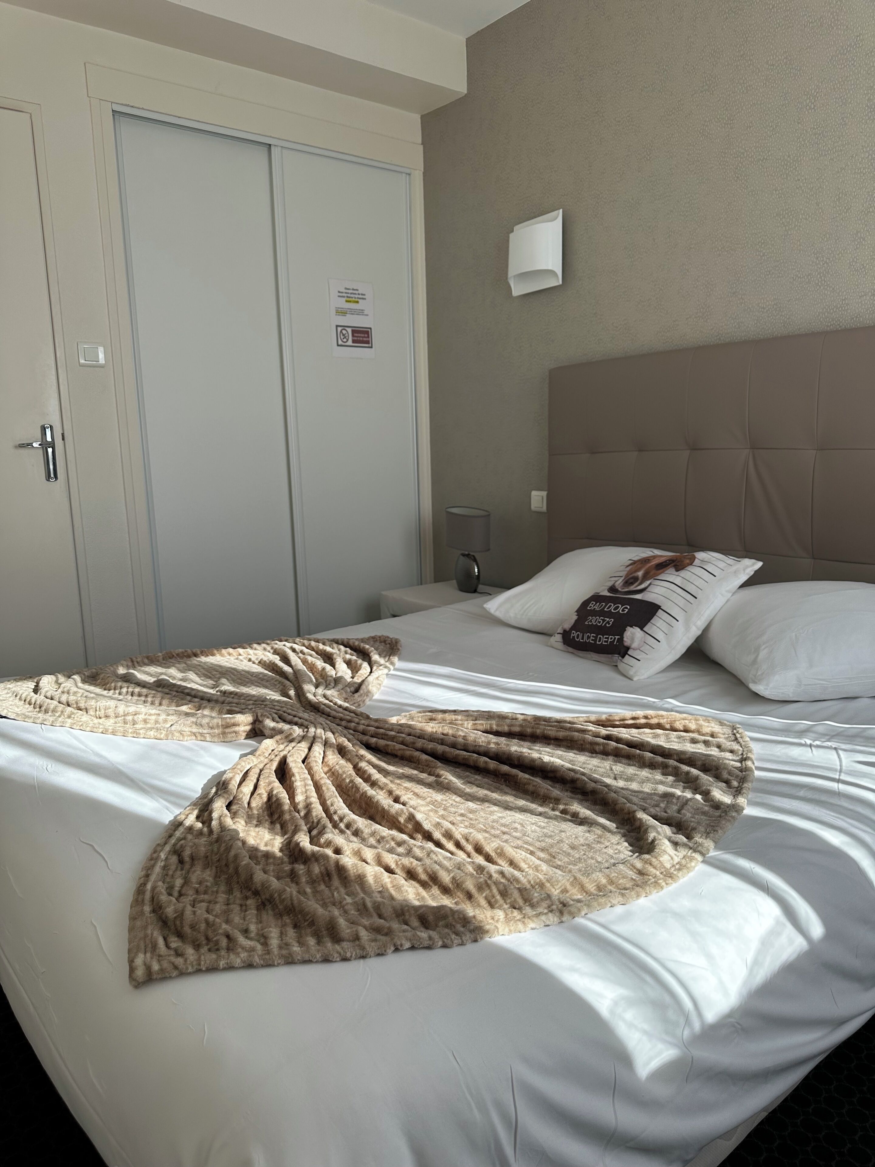 Quarto de hotel com cama, manta, travesseiro com pingente de coleira e mesa de cabeceira, além de luminária fixada na parede.