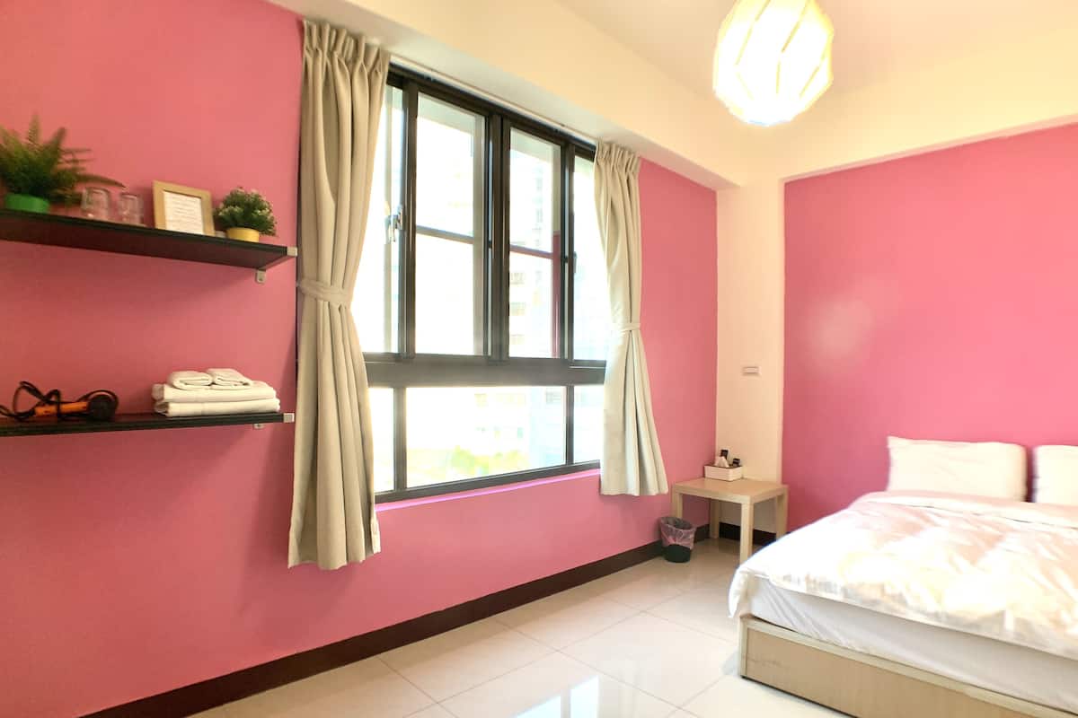 A bedroom with pink walls, a bed, a window with curtains, a small bedside table, and a shelf with items.