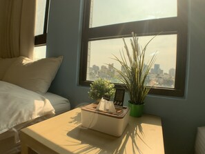 A hotel room with a bed, a bedside table with a tissue box, a potted plant, and a view of a cityscape through a window. - Partyport-Hai (Tainan)