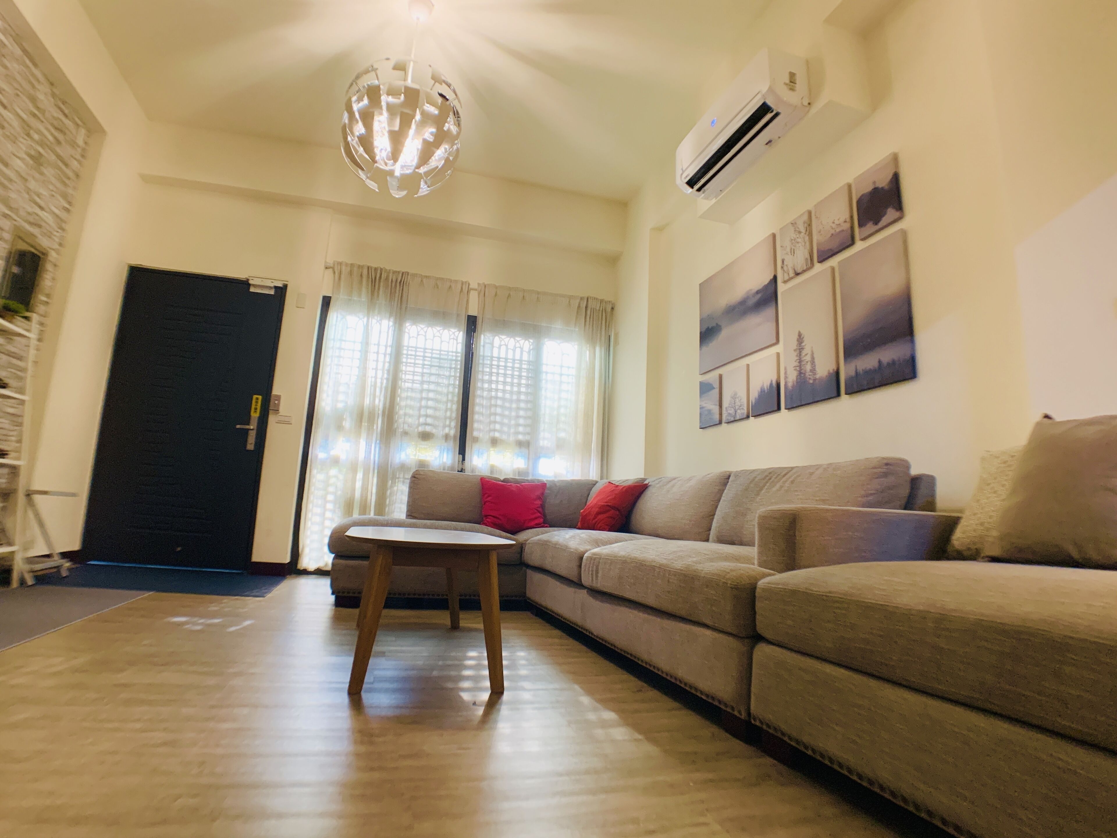 A living room with a sofa, a small table, and a chandelier. There is an air conditioning unit on the wall and a door in the background.