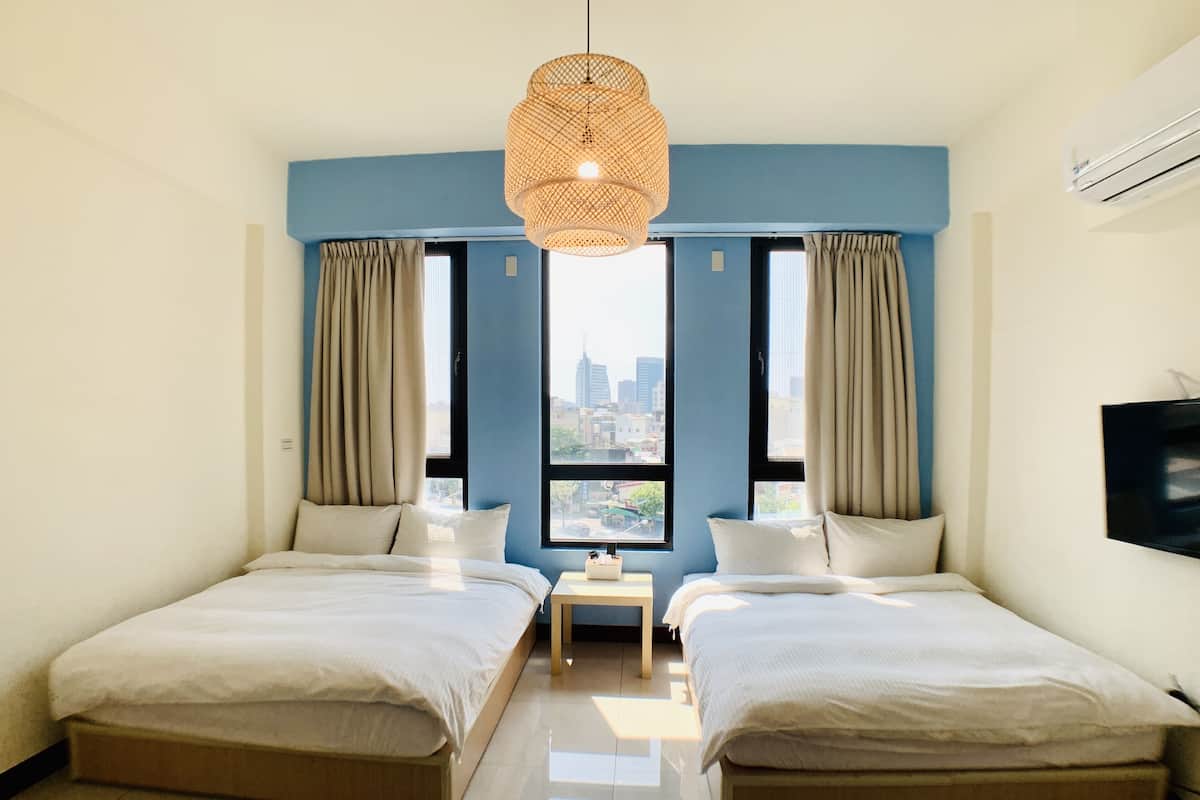 A modern hotel room with two beds, a city view, a hanging light fixture, and an air conditioning unit.