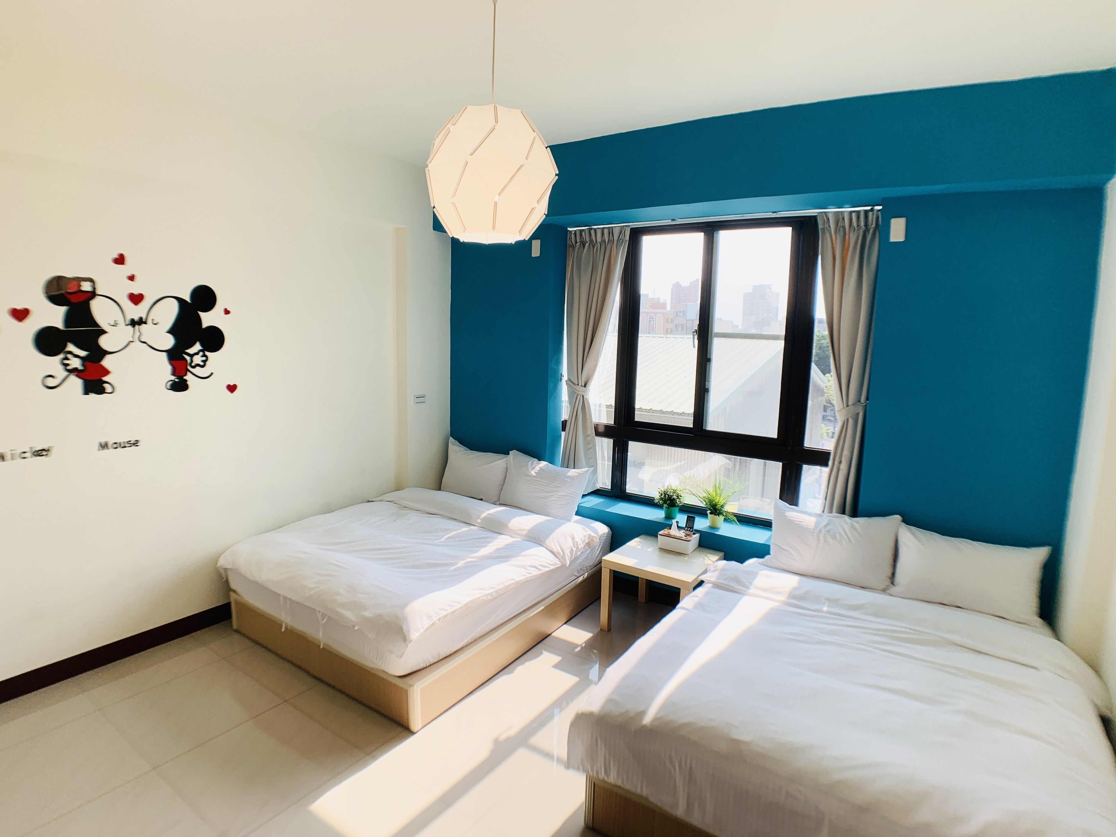 Hotelkamer met twee bedden, een raam met gordijnen, een tafeltje met een plant en een muurdecoratie van Mickey en Minnie Mouse.