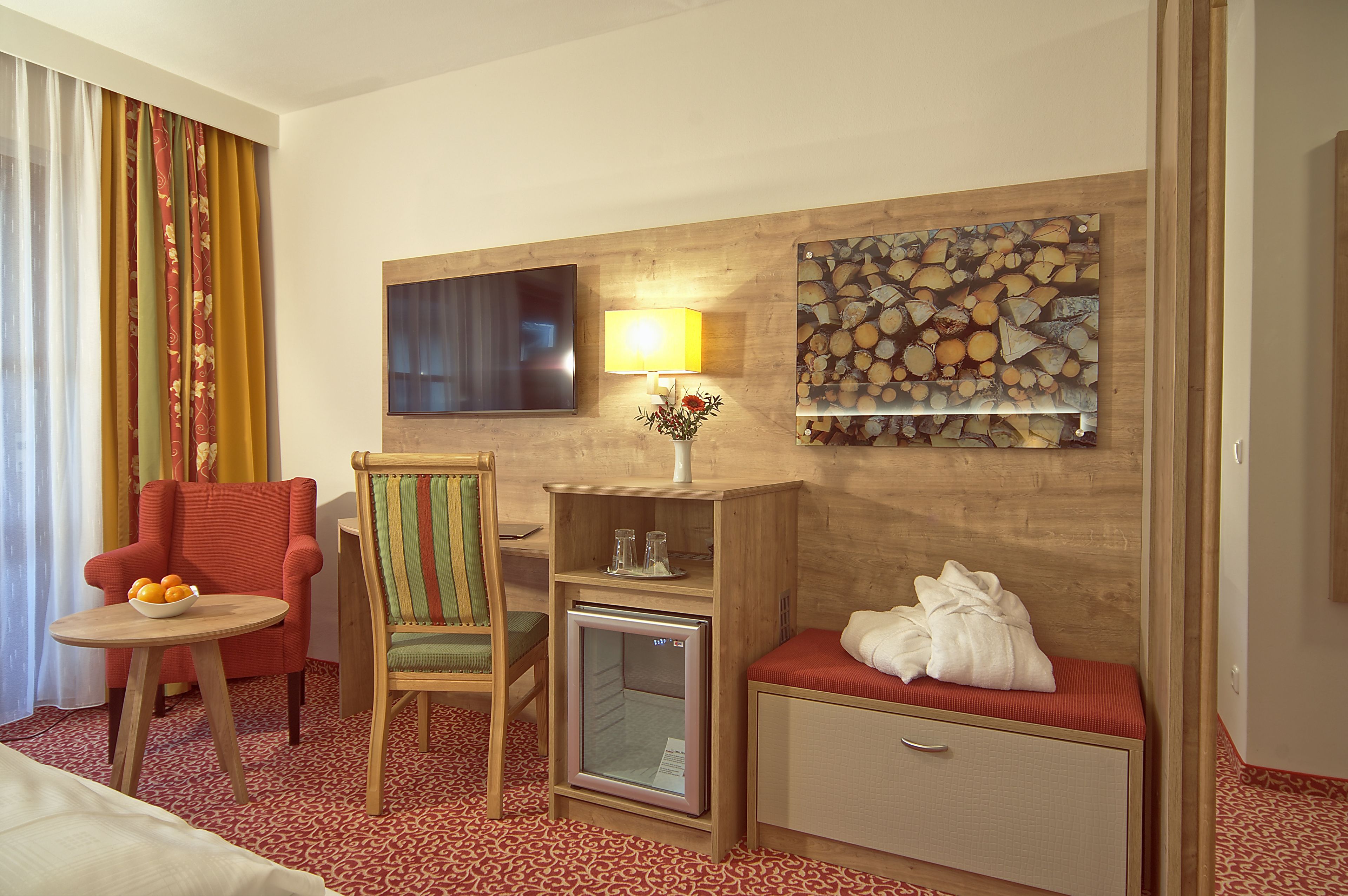 A hotel room with a bed, a red armchair, a wooden side table with a bowl of fruit, a small refrigerator, a lamp, and a wall-mounted TV.