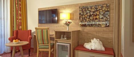 A hotel room with a bed, a red armchair, a wooden side table with a bowl of fruit, a small refrigerator, a lamp, and a wall-mounted TV.