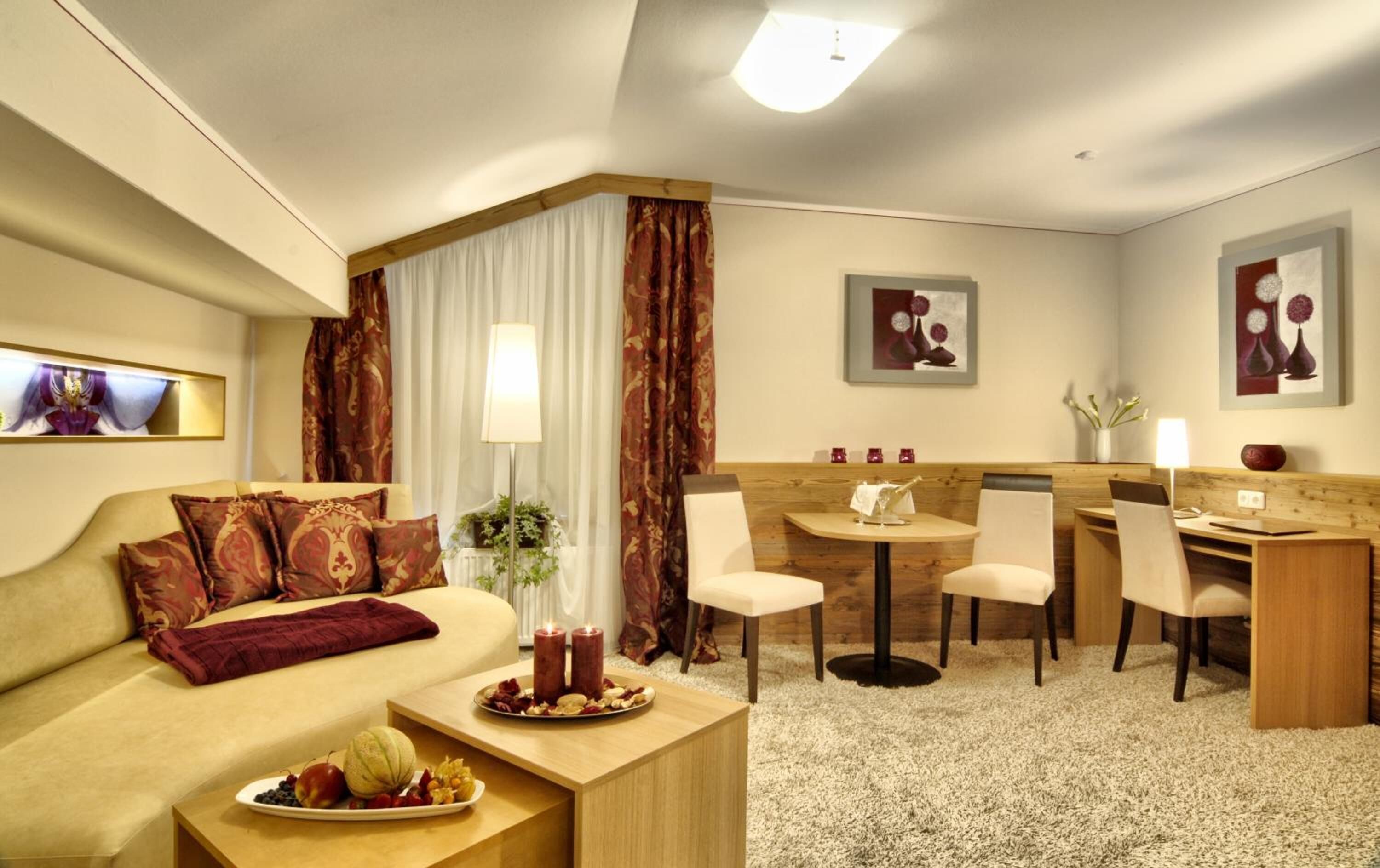 A hotel room with a sofa, a dining table with chairs, a small kitchen area, and a large window with curtains.