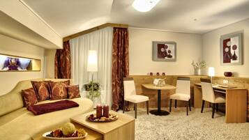 A hotel room with a sofa, a dining table with chairs, a small kitchen area, and a large window with curtains.