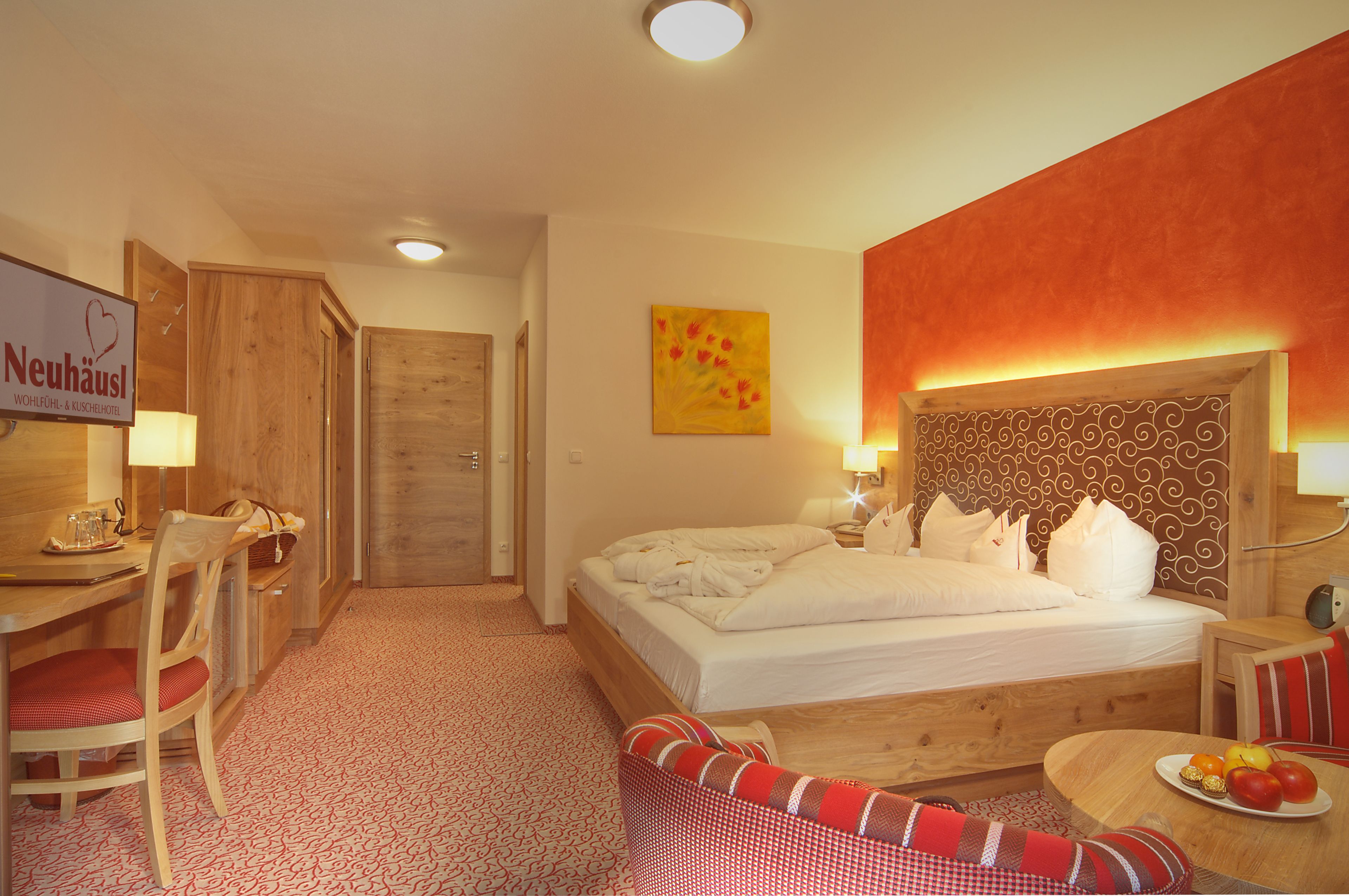 Une chambre d’hôtel avec un lit, un bureau, une chaise, une table à manger avec des fruits et une enseigne Neuhaus.