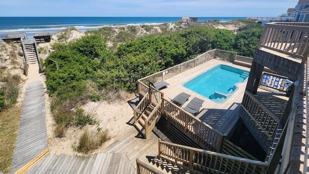 A wooden deck with a pool and hot tub, surrounded by greenery and overlooking the beach.