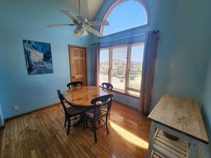 A dining area with a round wooden table and chairs, a ceiling fan, a window with blinds, and a painting on the wall.