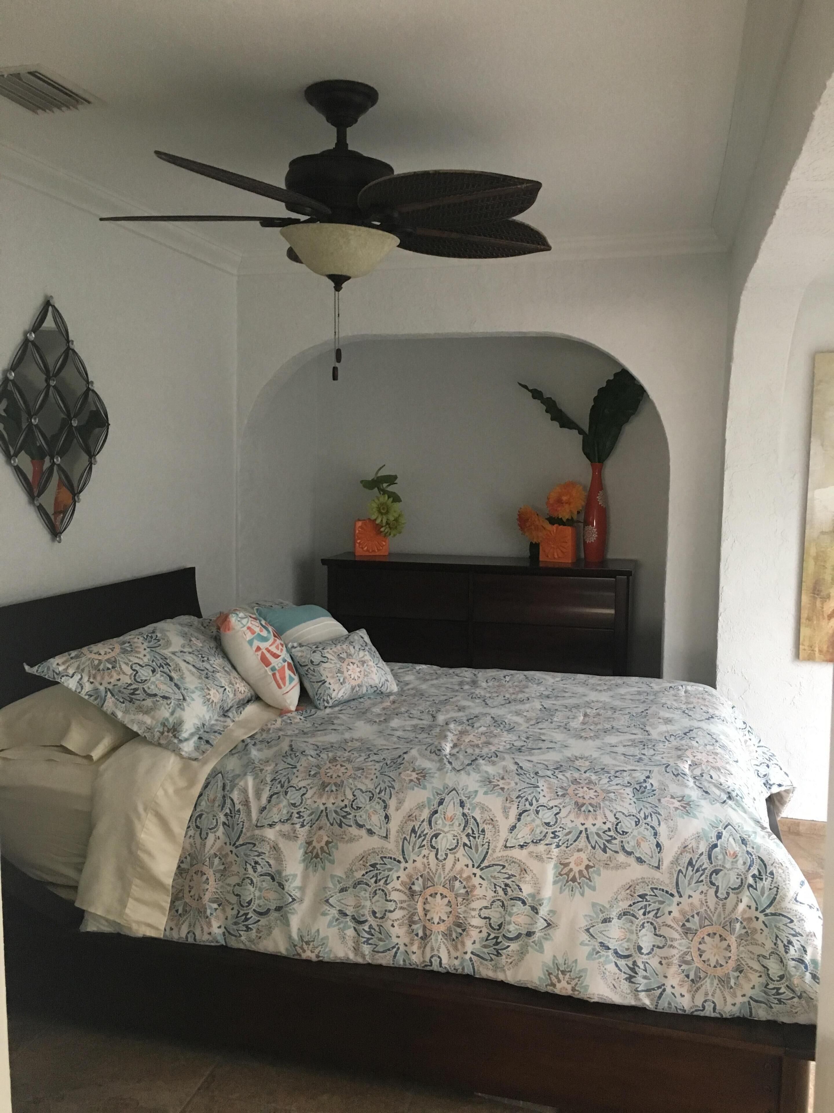 A bedroom with a bed, a ceiling fan, a decorative wall piece, a nightstand with a vase, and a painting on the wall.