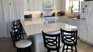Uma cozinha moderna com armários brancos, uma ilha central com banquetas, micro-ondas, fogão e geladeira decorada com imãs.