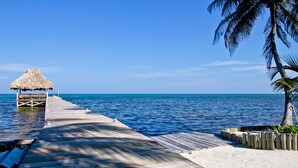 Eine Holzstege führt zu einer mit Stroh gedeckten Struktur am Meer, mit einer Palme und einer Sandfläche in der Nähe.