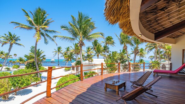 Eine Holzterrasse mit Liegestühlen und einem Tisch, mit Blick auf einen Strand mit Palmen und dem Meer.