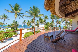 Eine Holzterrasse mit Liegestühlen und einem Tisch, mit Blick auf einen Strand mit Palmen und dem Meer.