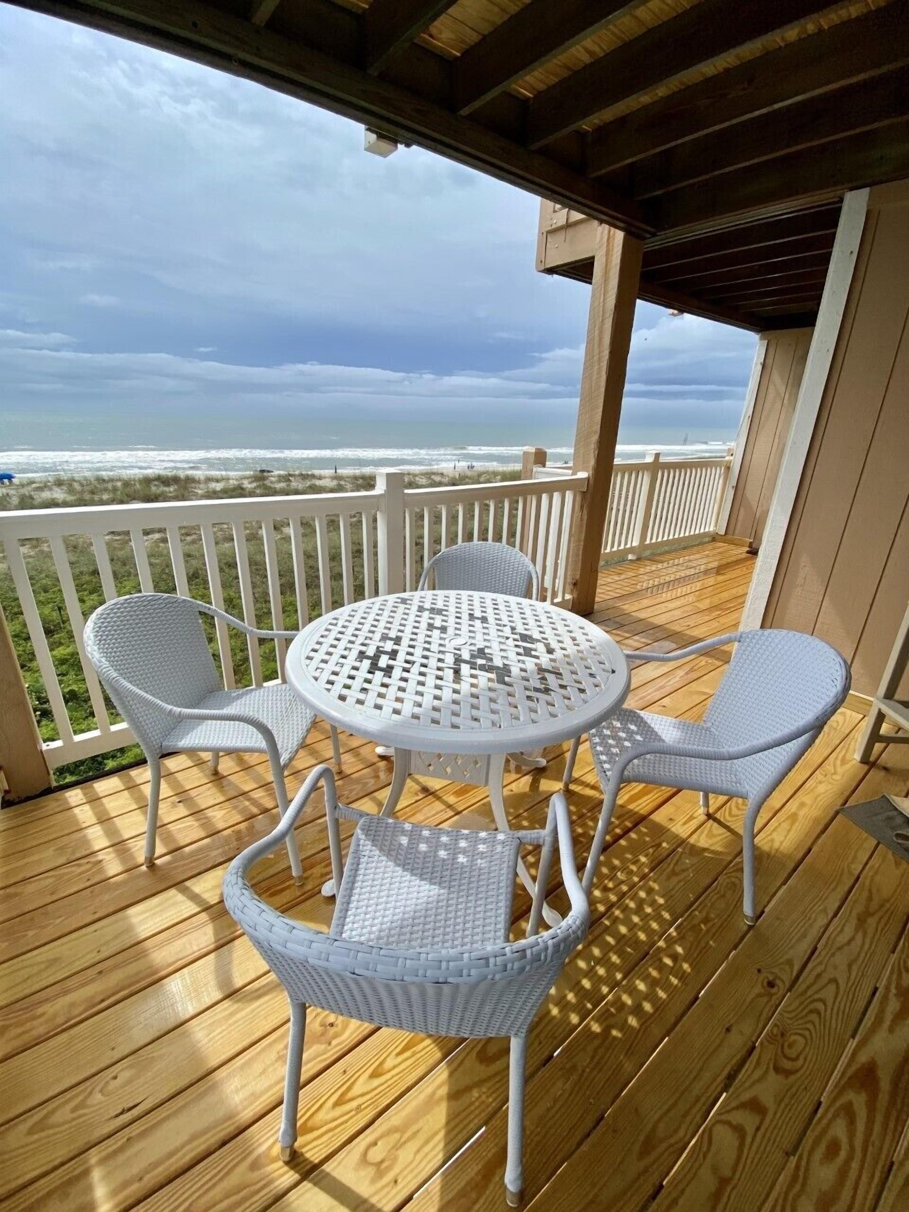Una terraza de madera con una mesa metálica blanca y cuatro sillas de mimbre, con vista a una playa.