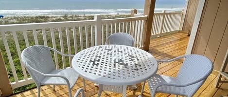 A wooden deck with a white metal table and four wicker chairs, overlooking a beach.
