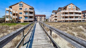 Un paseo marítimo de madera que lleva a un complejo de apartamentos frente al mar, con varios balcones y un cielo despejado.