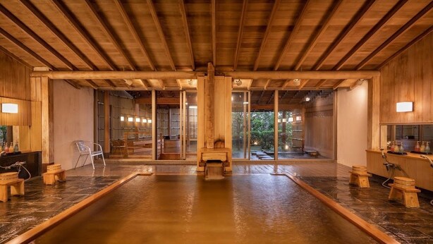 Un onsen japonés tradicional con vigas de madera, una fuente termal central y taburetes de madera.