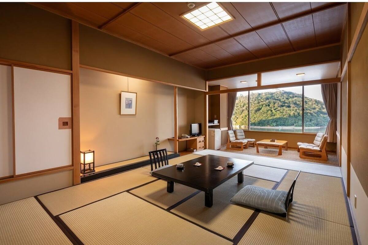 A traditional Japanese room with tatami flooring, a low table, and a view of a forested hill.
