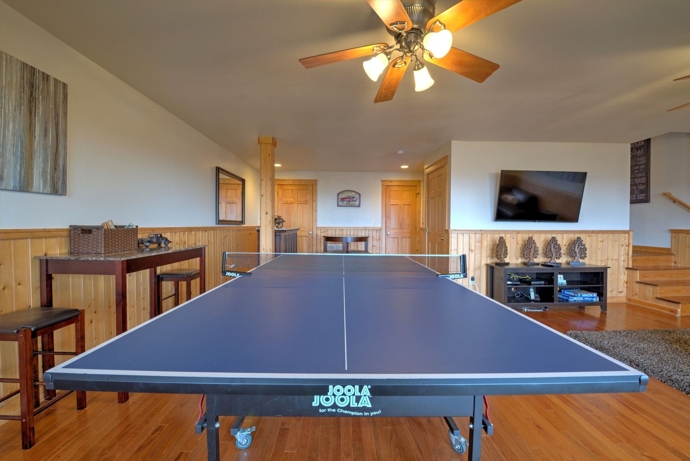 A ping pong table in a room with wooden floors and walls, a ceiling fan, and a television on the wall.