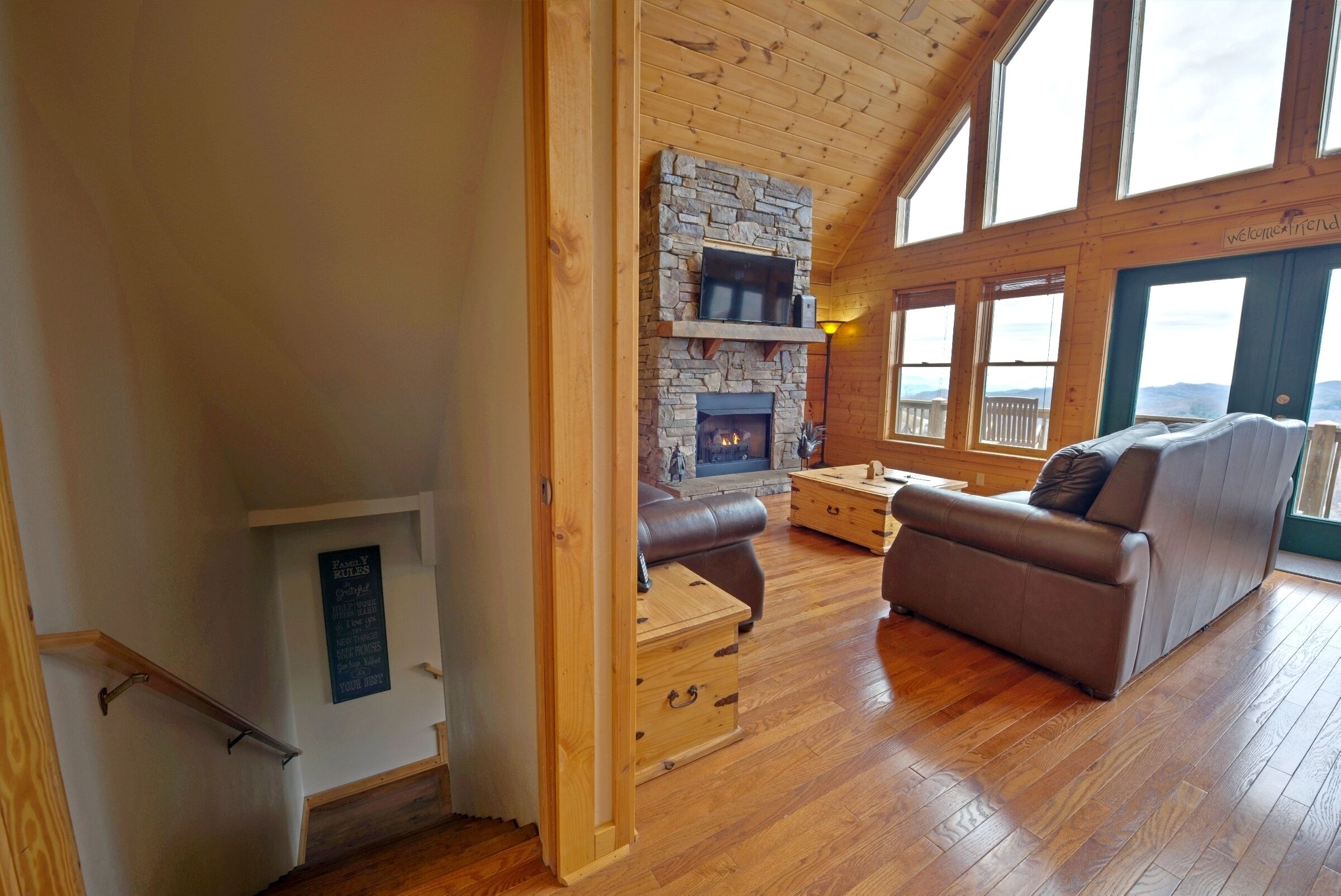 A cozy living room with a stone fireplace, wooden flooring, and a large window overlooking a deck.