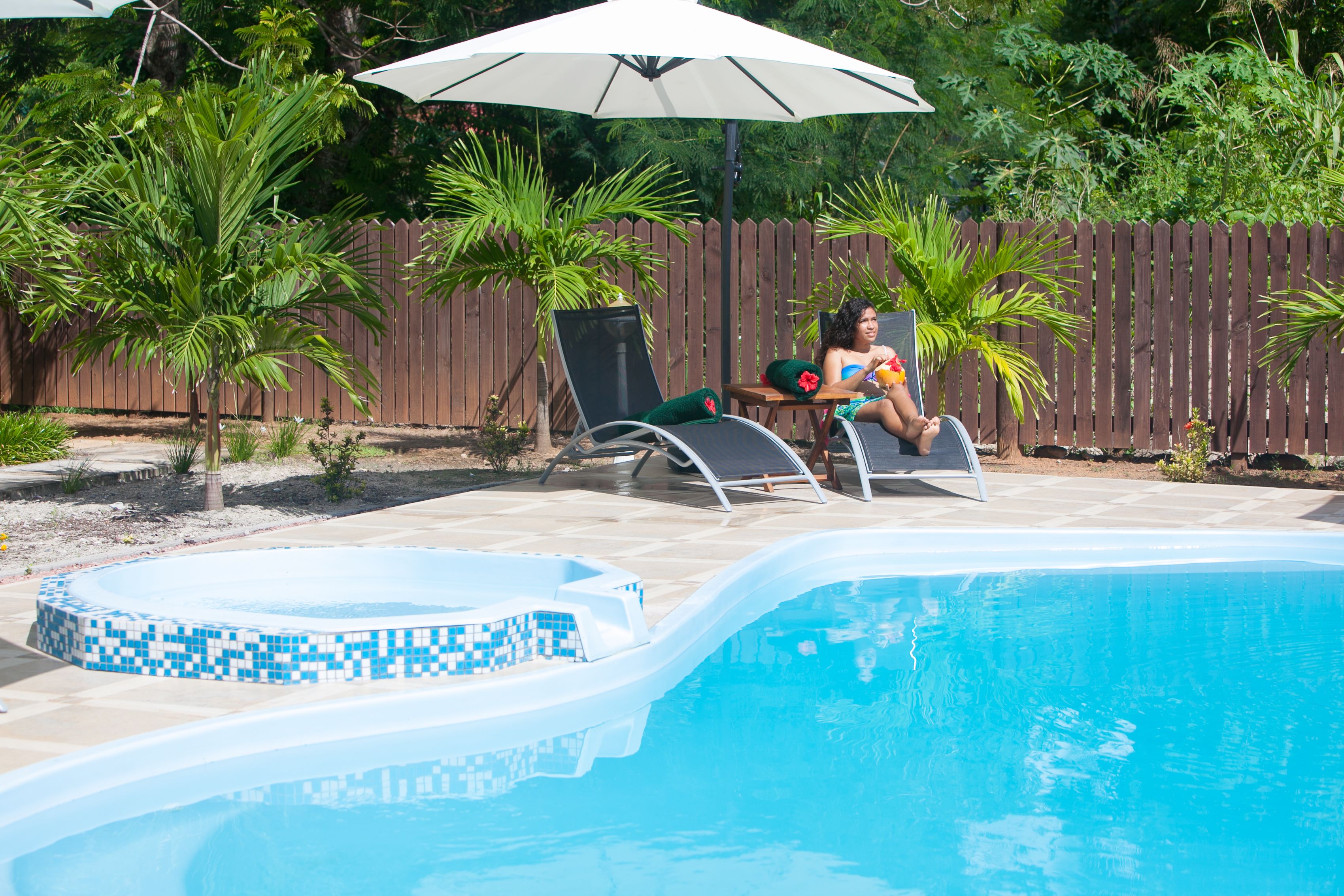 A swimming pool with a tiled edge, a lounge chair, and a small table with a person sitting.