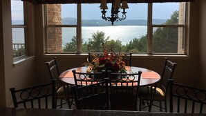 A dining area with a table and chairs, a chandelier, and a view of a lake through large windows. - Relax In Luxury On Table Rock Lake, Close To Branson (Hollister)