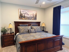 A bedroom with a wooden bed, two bedside tables with lamps, a nightstand, a potted plant, and a painting of a mountain landscape.