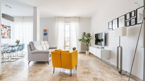 Ein modernes Wohnzimmer mit einer Couch, einem gelben Sessel, einem Essbereich mit einem Glastisch und einem an der Wand montierten Fernseher.