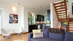 Una sala moderna con un sofá gris, dos sillas blancas, una escalera de madera y una cocina con armarios oscuros y un televisor.
