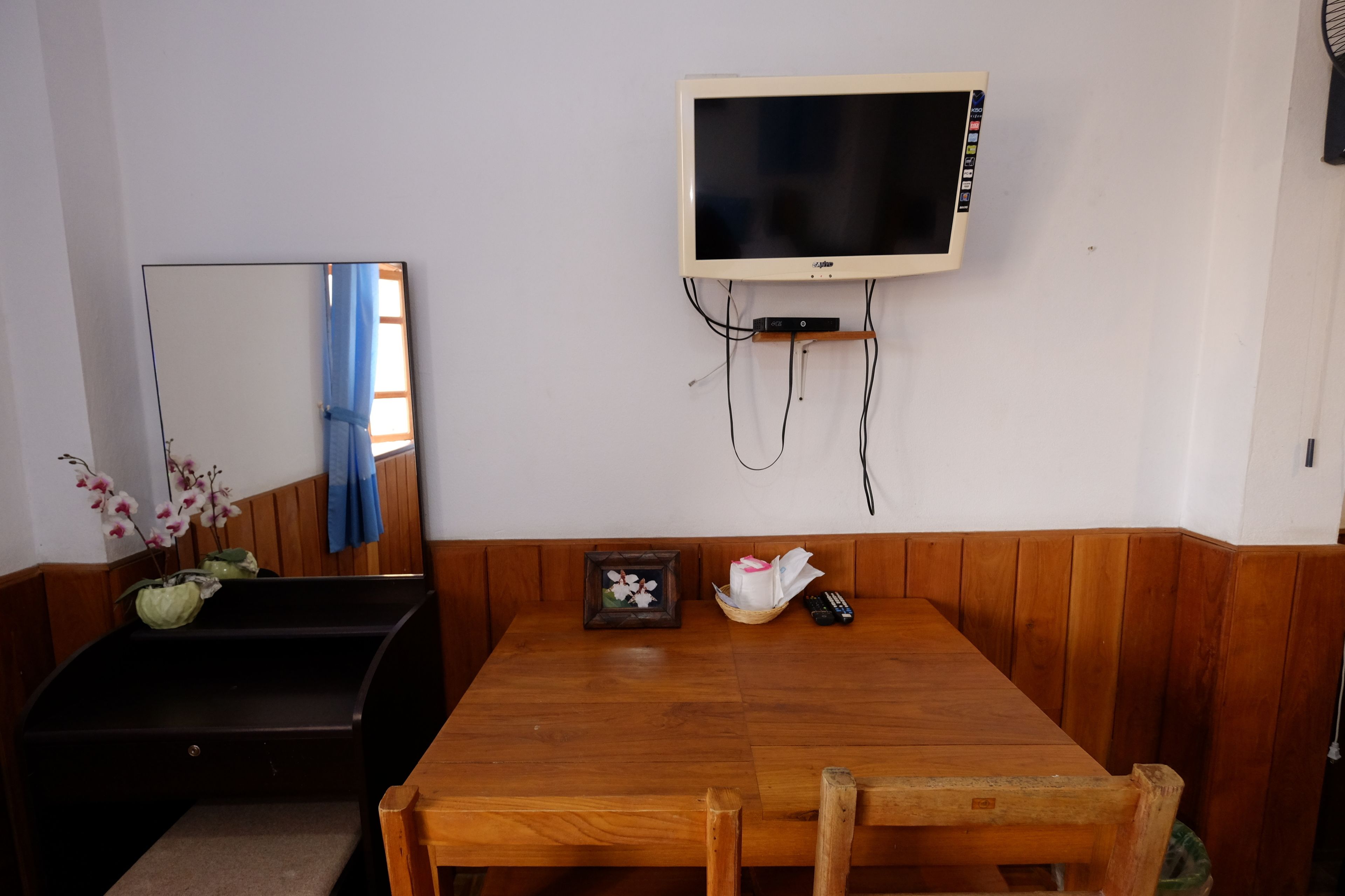A room with a wooden dining table, a television mounted on the wall, a mirror, and a small framed picture on the table.
