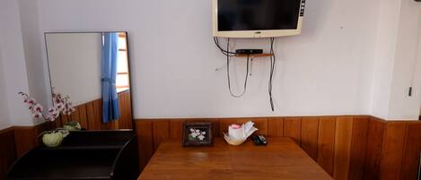 A room with a wooden dining table, a television mounted on the wall, a mirror, and a small framed picture on the table.