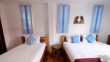 A hotel room with two single beds, wooden headboards, white bedding, blue curtains, and a small wooden stool.