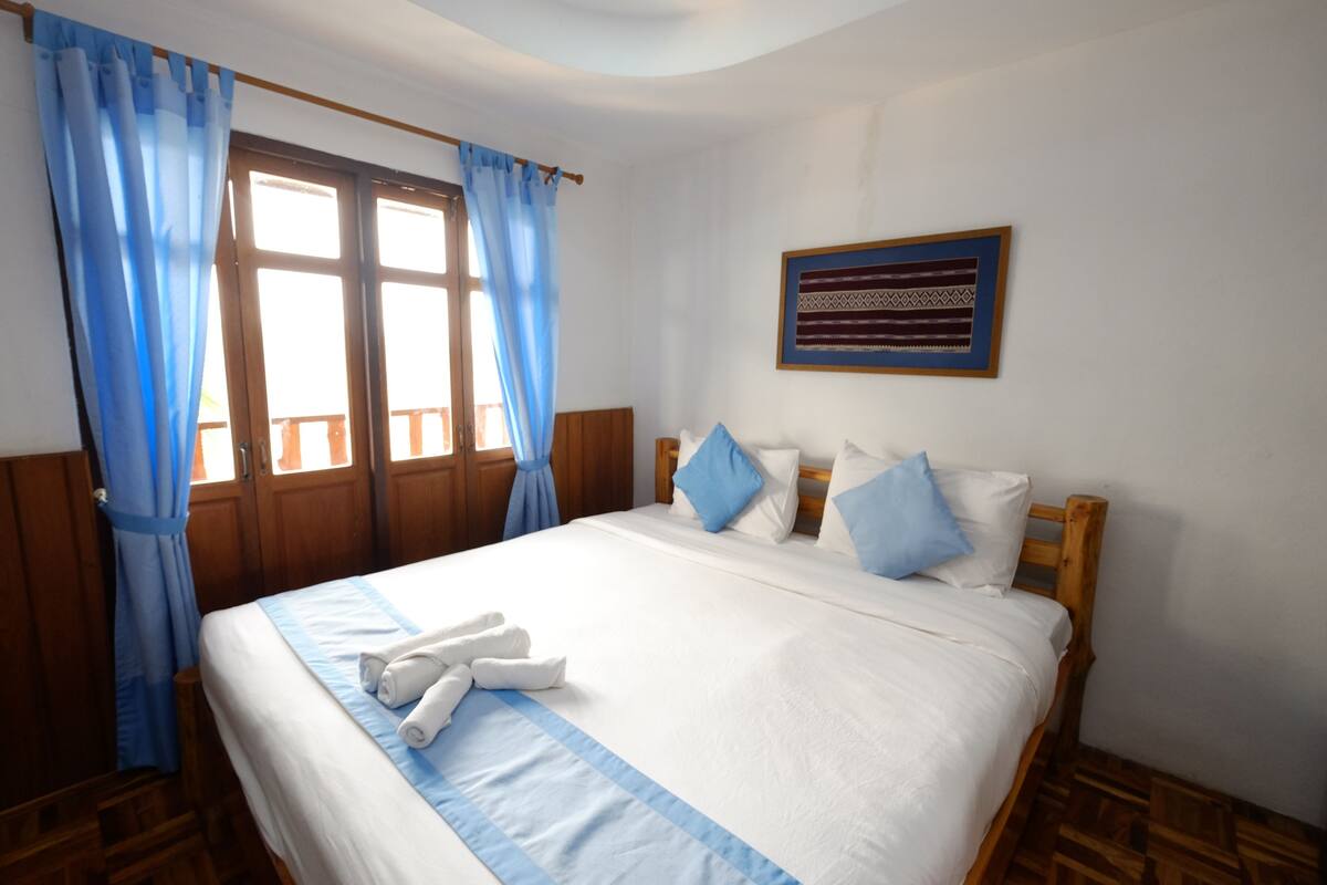 Uma cama bem arrumada com roupa de cama branca e azul, cabeceira de madeira e cortina listrada em azul.