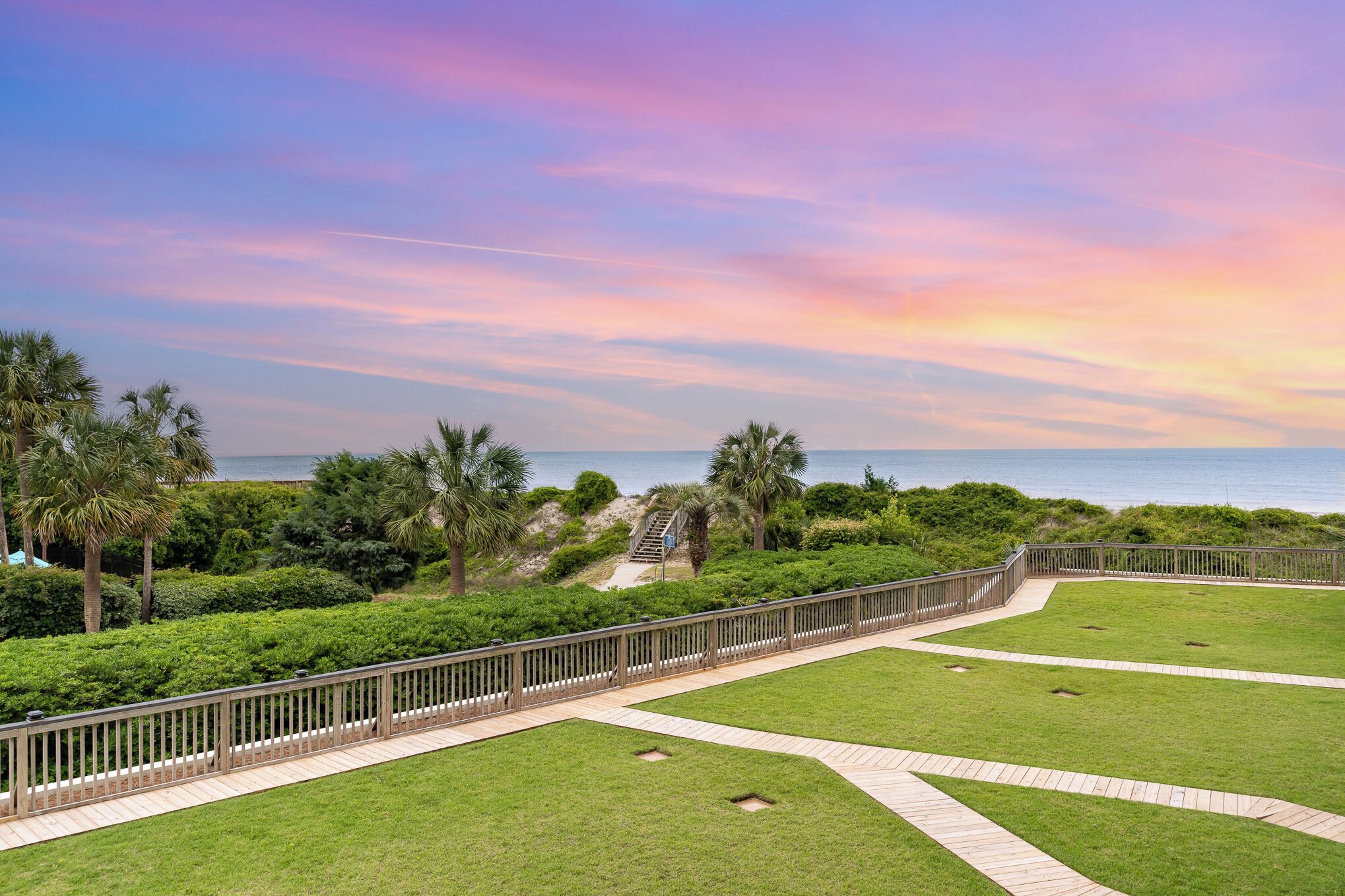 A landscaped area with a wooden pathway, palm trees, and a view of the sea.