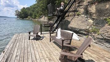 A wooden dock with Adirondack chairs and a bench, overlooking a calm lake with a rocky shoreline and trees.