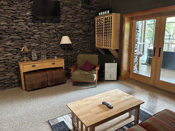 Una sala de estar acogedora con una pared de piedra, un mueble de madera con botellas de vino, un sillón cómodo, una mesita con un control remoto y una puerta corrediza de vidrio que da a un espacio al aire libre.