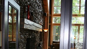 Une cheminée en pierre avec un manteau en bois, un plancher en bois et une vue sur l’extérieur grâce à de grandes fenêtres.