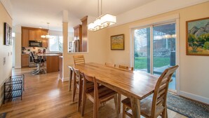 A spacious dining area with a wooden table and chairs, a kitchen in the background, and large windows offering a view of the outdoors. - Hazel House - Gorgeous, Views, Perfect Hood River Getaway! (Hood River)